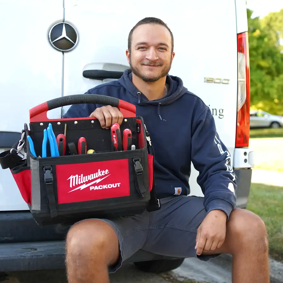 Plumber holding tools sitting on the bumper of a van